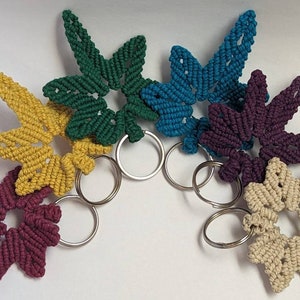 May include: Six colorful star-shaped keychains with silver key rings. The keychains are made of knotted string in shades of green, blue, purple, yellow, beige, and maroon.