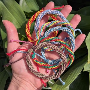 May include: A collection of colorful woven bracelets in various shades of red, yellow, blue, green, and brown. The bracelets are handmade with a braided design and adjustable ties. The bracelets are displayed against a backdrop of green foliage.
