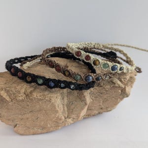 May include: Four handmade bracelets with different colored beads and natural fiber cords. The bracelets are stacked on a brown rock.