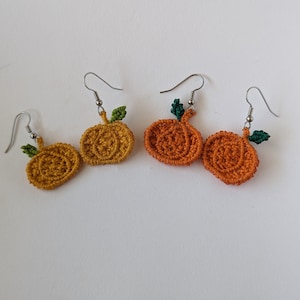 May include: A pair of crocheted pumpkin earrings with green leaves. The earrings are made with orange and yellow yarn and have silver hooks.