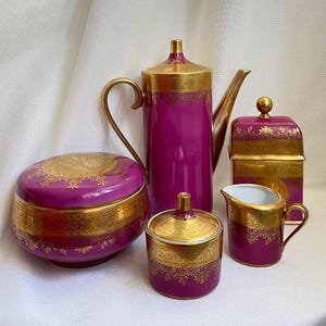 Könnte beinhalten: Ein fünfteiliges Porzellan-Teeservice in Lila und Gold. Das Set besteht aus einer Teekanne, einer Zuckerdose, einem Sahnekännchen, einem Deckelgefäß und einer rechteckigen Box. Jedes Teil hat goldene Verzierungen und dekorative Muster.