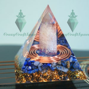 Könnte beinhalten: Eine klare Kristallpyramide mit einer Kupferspule und blauen Lapislazuli-Steinen. Die Pyramide steht auf einer Holzoberfläche.