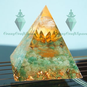 May include: A green and gold crystal pyramid with a clear quartz crystal at the top and a gold lotus flower inside. The pyramid is sitting on a black surface with gold lines.