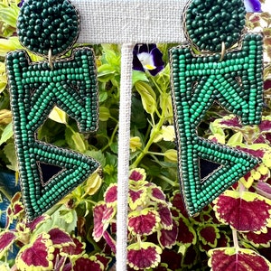 May include: A pair of green and gold geometric beaded earrings. The earrings are shaped like a triangle with a circle at the top. The earrings are hanging from a white stand.