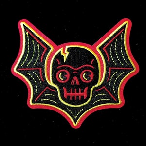 May include: A black and yellow embroidered patch with a skull and bat wings. The skull has a lightning bolt crack across the forehead and large, round eyes. The wings are outlined in red.