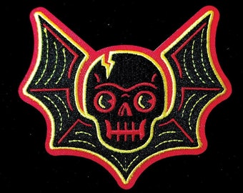 May include: A black and yellow embroidered patch with a skull and bat wings. The skull has a lightning bolt crack across the forehead and large, round eyes. The wings are outlined in red.