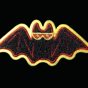 May include: A black embroidered bat with red stitching and yellow outline wearing sunglasses. The bat is a patch with a fuzzy texture.
