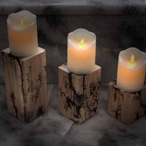 Puede incluir: Tres velas blancas en forma de pilar con llamas parpadeantes se colocan sobre candelabros de madera. Los candelabros están hechos de madera natural con un acabado único, agrietado y marrón oscuro.