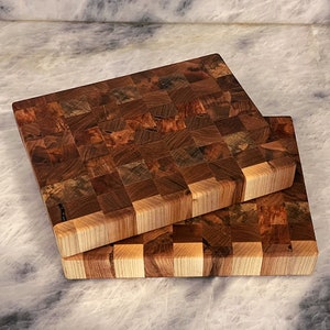 May include: Two wooden cutting boards stacked on top of each other. The top board is made of a dark wood with a checkered pattern. The bottom board is made of a lighter wood with a similar checkered pattern.