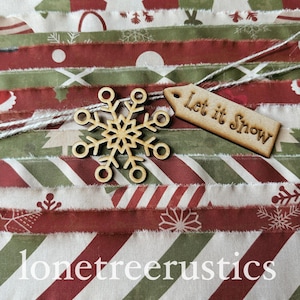 May include: A collection of fabric strips featuring red, green, and white holiday patterns including snowflakes and stripes. A wooden snowflake ornament and a wooden tag with the text Let it Snow are tied together with white twine.