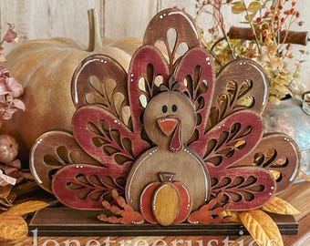 Puede incluir: Porta servilletas de madera en forma de pavo con un esquema de color rojo, marrón y naranja. El pavo tiene un diseño tallado y sostiene una pequeña calabaza. La base del porta servilletas es marrón y tiene el texto "lonetreerustrics" en él.