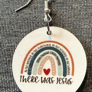 May include: A round white earring with a silver hook featuring a multi-colored rainbow graphic. The rainbow includes text that reads In the waiting in the searching in the healing in the hurting every minute every moment and a red heart. The text There was Jesus is printed below the rainbow.