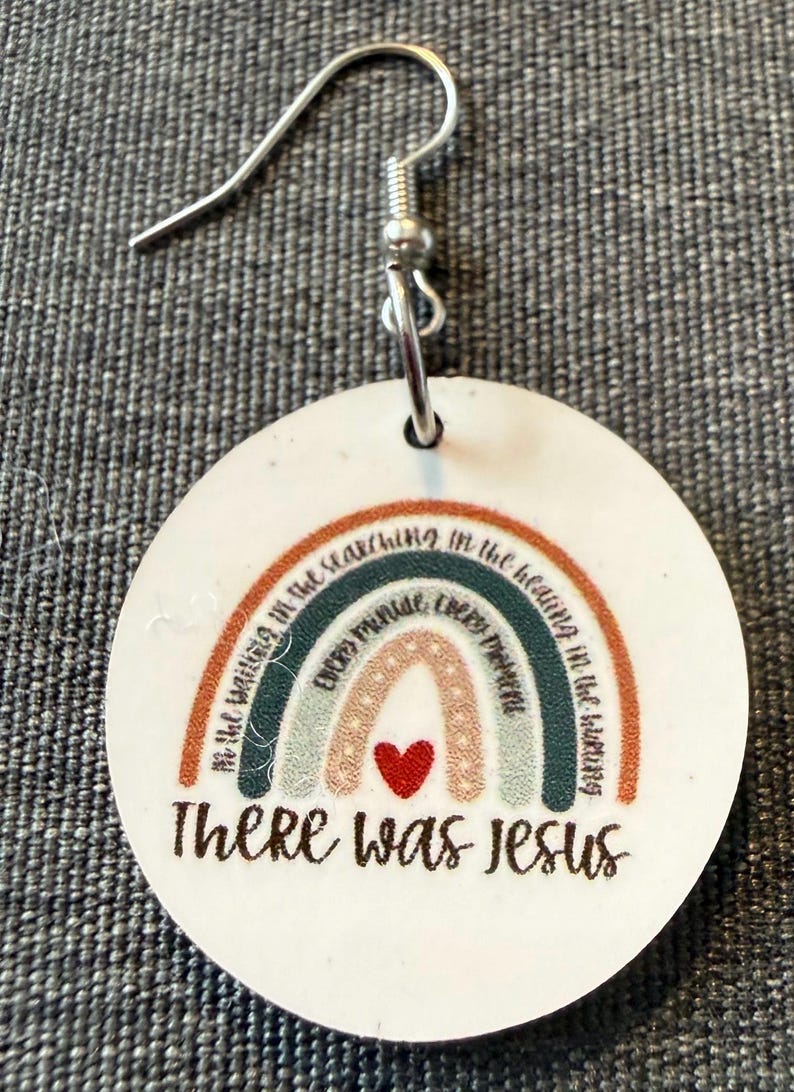 May include: A round white earring with a silver hook featuring a multi-colored rainbow graphic. The rainbow includes text that reads In the waiting in the searching in the healing in the hurting every minute every moment and a red heart. The text There was Jesus is printed below the rainbow.