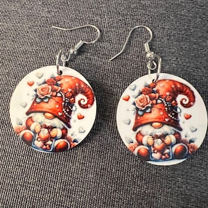 May include: A pair of earrings featuring a round, white background with a gnome holding a heart-shaped box. The gnome is wearing a red hat with white flowers and a white beard. The earrings are silver.