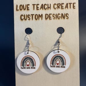 May include: A pair of round, white, flat earrings featuring a printed rainbow graphic with a small red heart at the center and the text "There Was Jesus" below. The earrings hang from silver-toned metal hooks on a light wood display card.