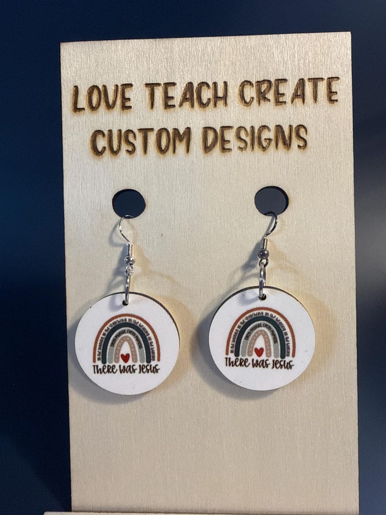 May include: A pair of round, white, flat earrings featuring a printed rainbow graphic with a small red heart at the center and the text "There Was Jesus" below. The earrings hang from silver-toned metal hooks on a light wood display card.