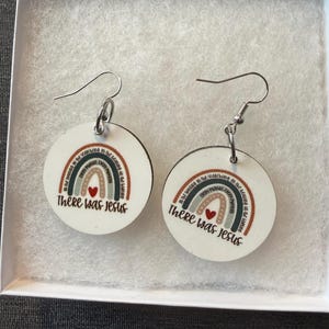 May include: A pair of round white dangle earrings featuring a printed rainbow graphic with a small red heart and the text "There was Jesus" written in black script. The earrings are attached to silver-toned metal hooks for pierced ears.