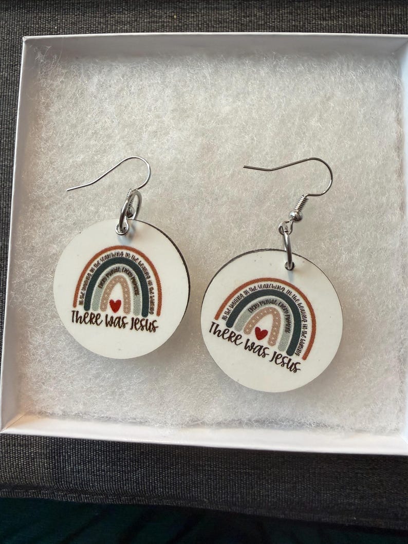 May include: A pair of round white dangle earrings featuring a printed rainbow graphic with a small red heart and the text "There was Jesus" written in black script. The earrings are attached to silver-toned metal hooks for pierced ears.