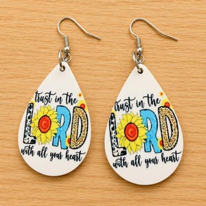 May include: Teardrop-shaped earrings featuring a white background with the text "trust in the LORD with all your heart" in black, blue, and leopard print lettering, accented by a yellow sunflower illustration and silver-toned metal hooks.