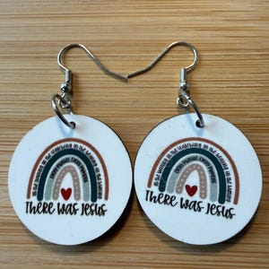 May include: A pair of round white dangle earrings featuring a printed rainbow design in muted earth tones. Each earring displays a small red heart and the text There Was Jesus, with additional small text arched along the rainbow stripes.