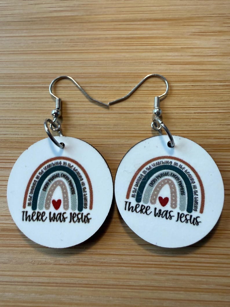 May include: A pair of round white dangle earrings featuring a printed rainbow design in muted earth tones. Each earring displays a small red heart and the text There Was Jesus, with additional small text arched along the rainbow stripes.