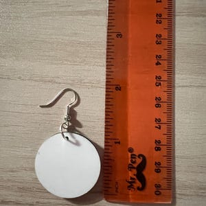 May include: A circular white earring with a silver-toned metal hook, positioned next to an orange ruler. The earring measures approximately 1.25 inches in diameter, as indicated by the markings on the ruler placed alongside the object.