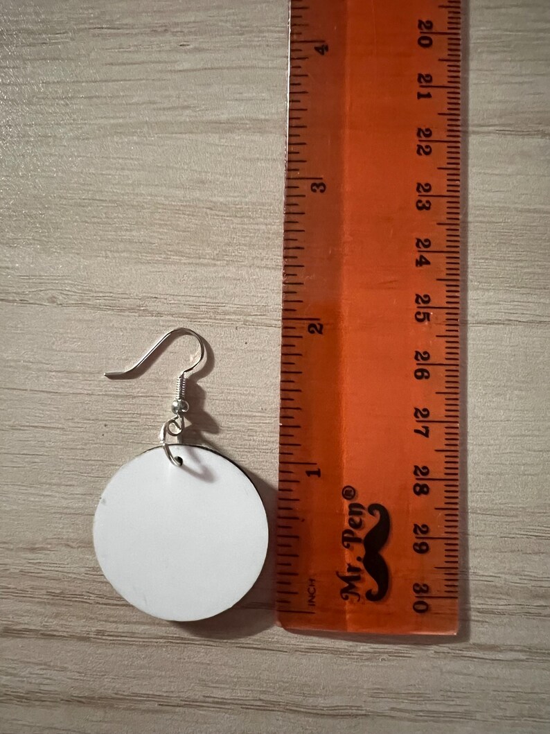 May include: A circular white earring with a silver-toned metal hook, positioned next to an orange ruler. The earring measures approximately 1.25 inches in diameter, as indicated by the markings on the ruler placed alongside the object.