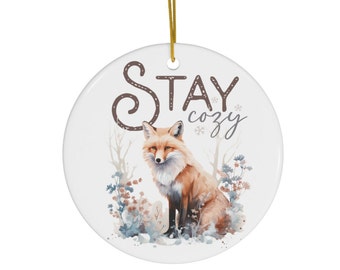 May include: A round ceramic ornament with a watercolor illustration of a fox sitting in a snowy forest. The ornament has the text "Stay cozy" in brown lettering.