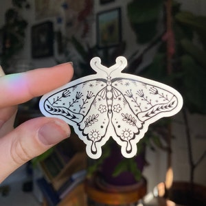 Peut inclure: Un autocollant transparent avec une illustration en noir et blanc d'un papillon avec des d&eacute;tails floraux. Le papillon a un corps en forme de c&oelig;ur et des motifs complexes sur ses ailes.