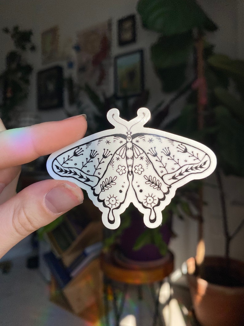 Peut inclure: Un autocollant transparent avec une illustration en noir et blanc d'un papillon avec des d&eacute;tails floraux. Le papillon a un corps en forme de c&oelig;ur et des motifs complexes sur ses ailes.