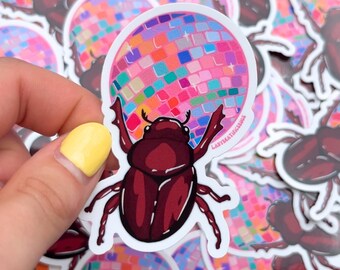 May include: A sticker featuring a cartoon beetle holding a pink disco ball with multi-colored mirrored tiles. The sticker has the text "Ladymathissick" on the bottom right corner.