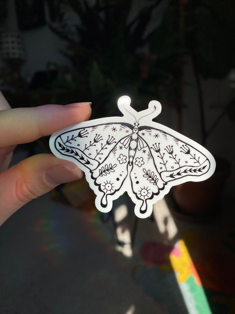Peut inclure: Un autocollant noir et blanc repr&eacute;sentant un papillon avec des d&eacute;tails floraux. Le papillon a un design stylis&eacute; avec des motifs complexes et une ambiance fantaisiste.