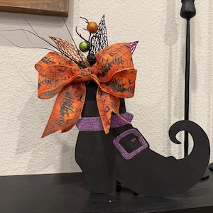 以下が含まれることがあります： クモの巣と「ハロウィン」の文字が入った大きなオレンジ色のリボンが付いた、黒い魔女のブーツの装飾。 ブーツには紫色のキラキラしたバンドとバックルが付いています。 装飾的な小枝、ベリー、黒いウェブが上部をアクセントにしています。
