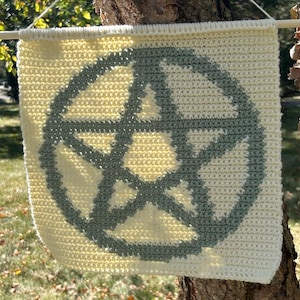 Könnte beinhalten: Ein cremefarbenes, gehäkeltes Wandbehang mit einem grauen Pentagramm-Design. Das Banner ist an einem Holzstab aufgehängt und hängt vor einem Baumstamm und grünem Gras. Das Design ist ein Symbol für Wicca und Paganismus.
