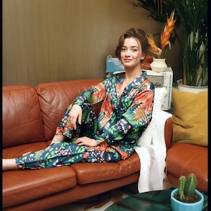 Puede incluir: Un conjunto de pijama con estampado floral en tonos verde azulado, naranja y verde. El conjunto incluye una camisa con botones y pantalones a juego. La persona est&aacute; sentada en un sof&aacute; de cuero marr&oacute;n. Un cactus en una maceta azul es visible.
