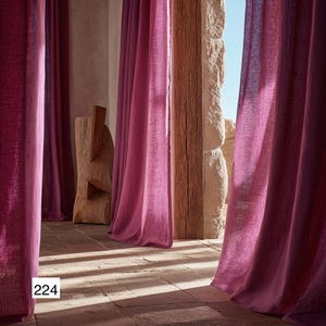 Op de afbeelding: Interieur met vloeiende, magenta gordijnen. De gordijnen omlijsten een uitzicht op een stenen muur en houten balken. De vloer is van stenen tegels. Het nummer "224" is zichtbaar in een wit kader linksonder.