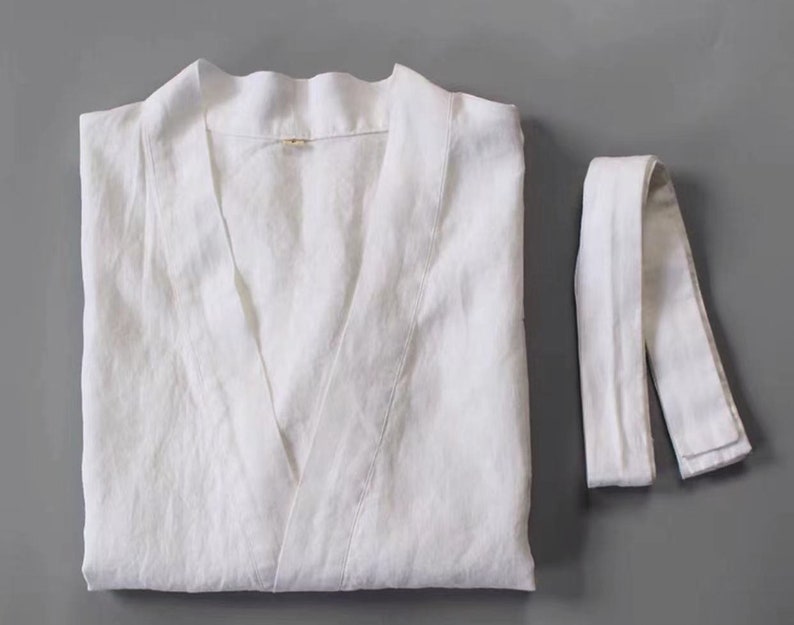 May include: White linen robe with a V-neck and a matching tie belt. The robe is folded and laid flat on a grey surface.