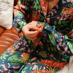 Puede incluir: Un conjunto de pijama de dos piezas con un estampado bot&aacute;nico y de tigre vibrante. El conjunto presenta un fondo verde azulado oscuro con dise&ntilde;os florales y de animales en naranja, rosa y verde. La parte superior de manga larga tiene botones.