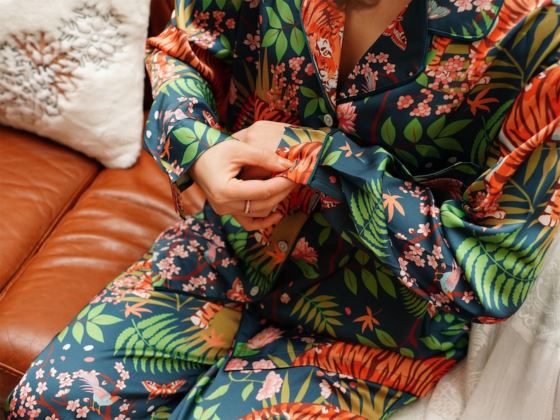 Puede incluir: Un conjunto de pijama de dos piezas con un estampado bot&aacute;nico y de tigre vibrante. El conjunto presenta un fondo verde azulado oscuro con dise&ntilde;os florales y de animales en naranja, rosa y verde. La parte superior de manga larga tiene botones.