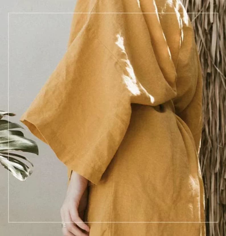 May include: A mustard yellow linen robe with long sleeves and a sash tie. The robe is made of a soft, lightweight fabric and has a relaxed fit.