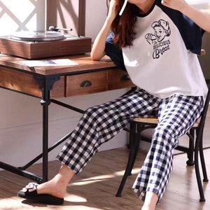May include: A woman wearing a white and blue checkered pajama pants with a white t-shirt featuring a cartoon illustration of a boy wearing a baseball cap and holding a loaf of bread. The t-shirt has the text "The Delicious Bread" printed on it.