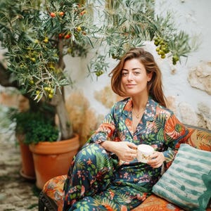 Puede incluir: Un conjunto de pijama con estampado floral en tonos verde azulado, naranja y verde. El conjunto incluye una camisa con botones y pantalones a juego. La persona sostiene una taza blanca y est&aacute; sentada en un banco estampado.