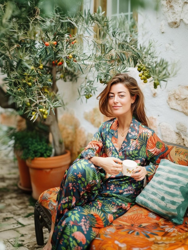 Puede incluir: Un conjunto de pijama con estampado floral en tonos verde azulado, naranja y verde. El conjunto incluye una camisa con botones y pantalones a juego. La persona sostiene una taza blanca y est&aacute; sentada en un banco estampado.