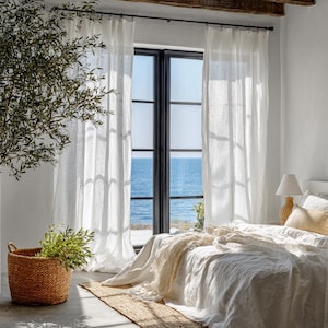 May include: A bright, airy bedroom with white linen curtains framing a view of the ocean. A bed with white bedding and a woven basket with greenery add to the room's natural aesthetic. A lamp sits on a bedside table.