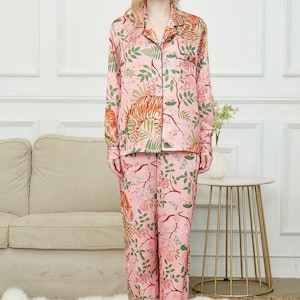 May include: Pink pajama set with a botanical and tiger print. The long-sleeved top has a collared neckline and button closure. The pants have a matching print. The set is trimmed with black piping.