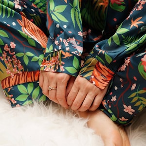 Puede incluir: Conjunto de pijama verde azulado oscuro con un estampado bot&aacute;nico vibrante con rayas de tigre naranjas, flores rosas y hojas verdes. La parte superior de manga larga y los pantalones son de material sedoso. El conjunto descansa sobre una superficie blanca y esponjosa.