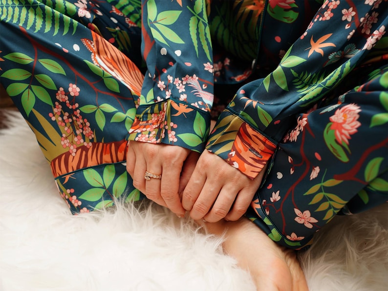 Puede incluir: Conjunto de pijama verde azulado oscuro con un estampado bot&aacute;nico vibrante con rayas de tigre naranjas, flores rosas y hojas verdes. La parte superior de manga larga y los pantalones son de material sedoso. El conjunto descansa sobre una superficie blanca y esponjosa.