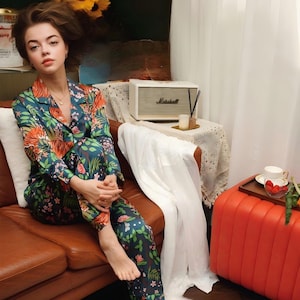 Puede incluir: Una persona con un conjunto de pijama con estampado floral en tonos verde azulado, naranja y rosa. El conjunto incluye una camisa con botones y pantalones a juego. La persona est&aacute; sentada en un sof&aacute; de cuero marr&oacute;n.