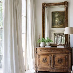 May include: An interior shot showing a detailed wooden cabinet with a large, ornate mirror. The mirror reflects a painting and a fireplace. White curtains frame a window, and a lamp stands nearby. The cabinet has carved details and is adorned with potted plants.