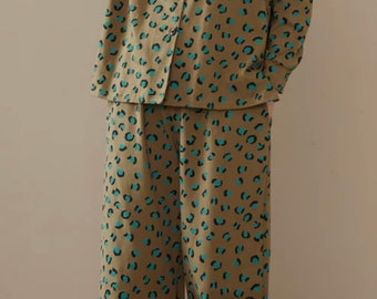 May include: A two-piece pajama set in a tan color with a leopard print pattern in teal and black. The top has a collared neckline and button-down front. The word "AKAY" is printed on the top. The pants are wide-legged.